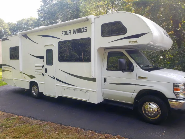 This clean white Class C motorhome, an ideal rv rental, is parked on an asphalt driveway with trees in background.. Thor Four Winds 2021