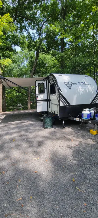 Peaceful RV rental setting. White travel trailer with awning extended at a green, tree-filled gravel campsite.. Heartland RVs Mallard 2022