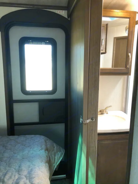 Inside this RV rental: A cozy bedroom with an exit door and a view of the compact bathroom sink.. Keystone RV Cougar 2021