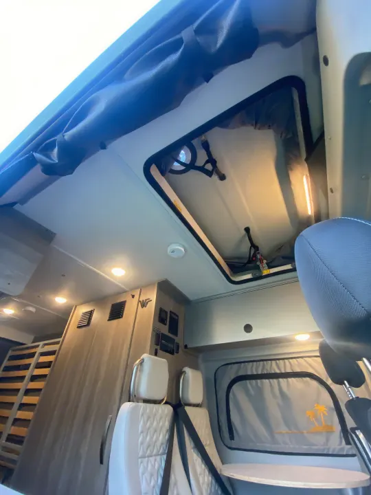 RV rental interior: Looking up at pop-top bed access, overhead lights, control panel, and front cabin seats.. Winnebago Solis 2023
