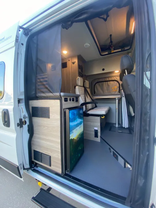 Inside a white RV rental, revealing a compact kitchen, portable fridge, versatile seating, and mesh screen door.. Winnebago Solis 2023