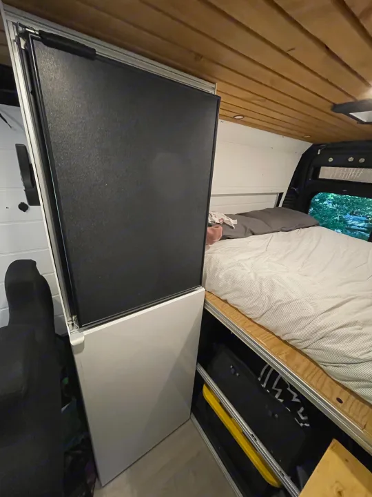 RV rental interior: A bed with striped bedding, upright fridge, and under-bed storage in a wood-paneled van.. RAM Promaster 2022