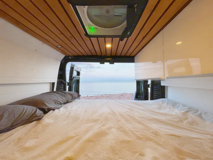 Cozy interior of an RV rental: a comfortable bed with striped sheets views a tranquil ocean and rocky beach at dusk.. RAM Promaster 2022