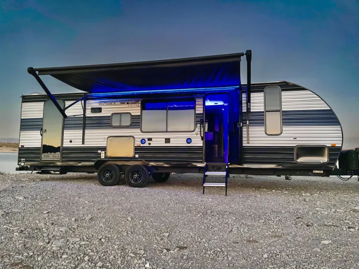 Modern RV rental: a travel trailer with bright blue LED lights & extended awning, parked on gravel for a night of camping.. Forest River Cherokee Grey Wolf 2022