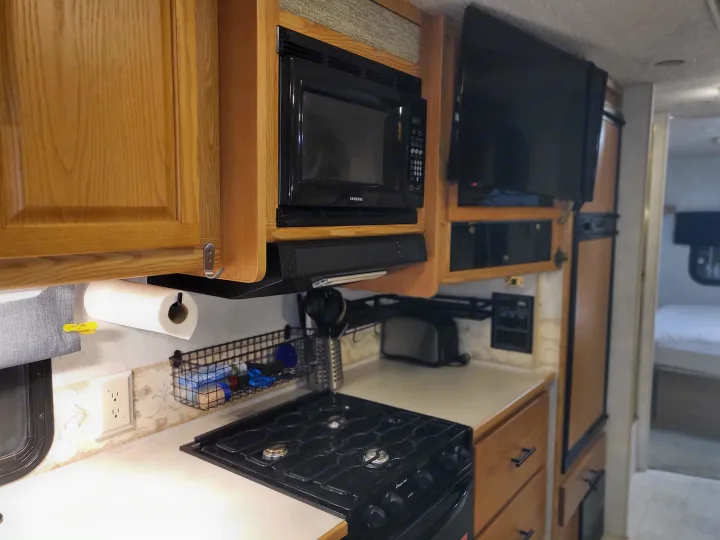 Compact RV rental kitchen with oak cabinets, microwave, TV, stovetop, fridge, and a paper towel holder. View to bedroom.. Itasca Spirit 2003