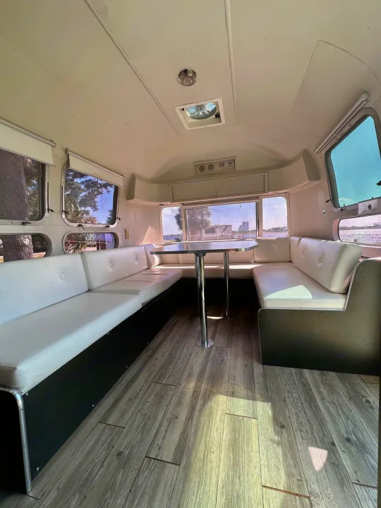 Clean Airstream RV rental interior: white cushioned dinette, central table, wood floor, and windows offering views.. Airstream Sovereign 1973