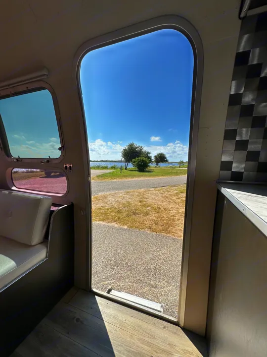 Experience nature with an RV rental! Open door view from inside an RV: tranquil lake, trees, and sunny blue sky.. Airstream Sovereign 1973
