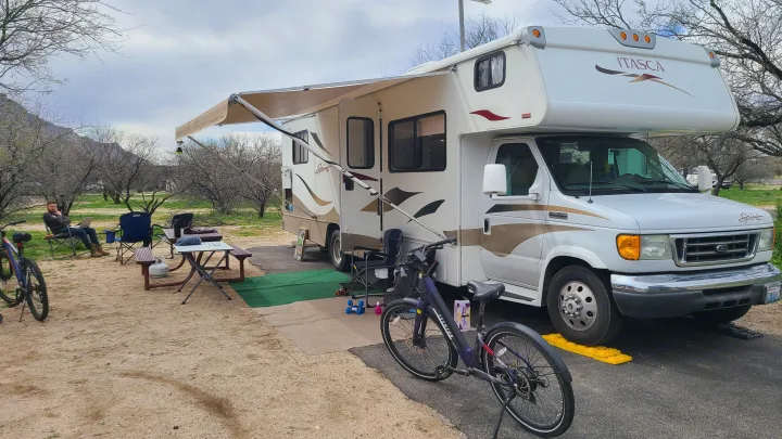 RV rental parked at a campsite with awning extended. A man uses a laptop, surrounded by bikes and camping gear.. Itasca Spirit 2007