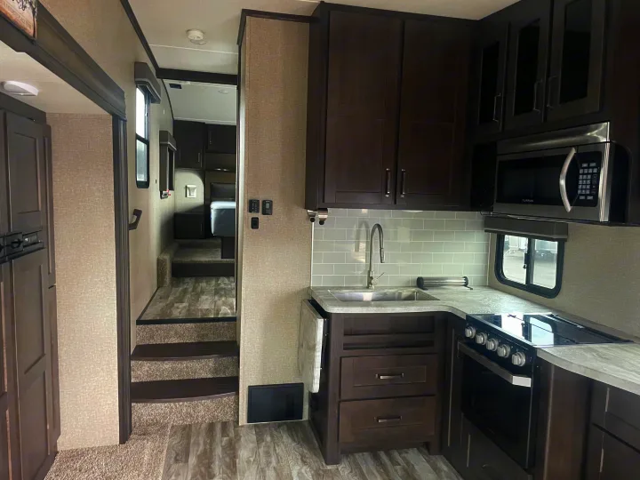 Modern RV rental interior showing a kitchen with dark cabinets, microwave, and stove; steps lead to a raised area.. Grand Design Reflection 2021