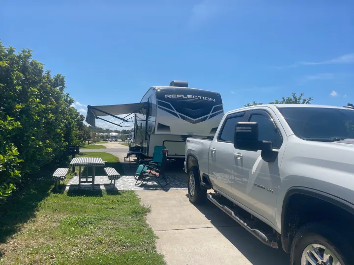 An RV rental (Reflection travel trailer) with its awning extended is parked alongside a white pickup truck at a sunny campground site, next to a picnic area.. Grand Design Reflection 2021