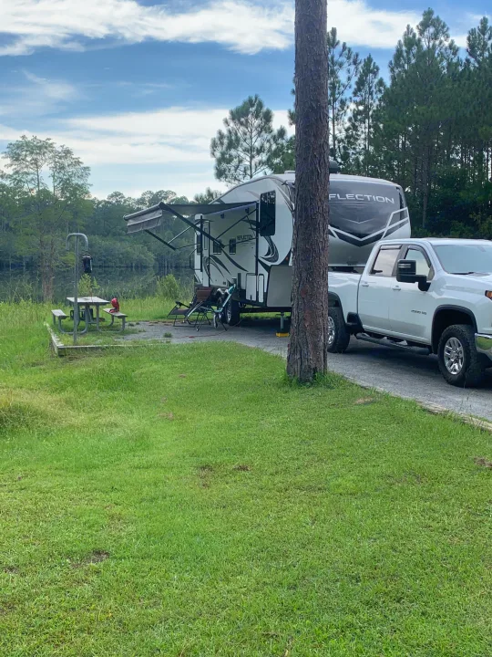 A white fifth-wheel RV and pickup truck at a lakeside rv rental campsite, with extended awning, picnic table, and chairs.. Grand Design Reflection 2021