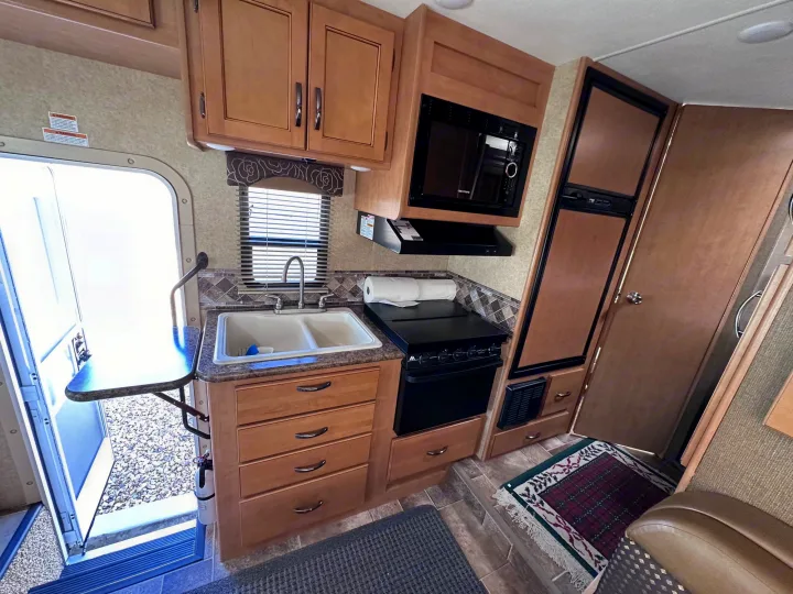 This RV rental interior showcases a full kitchen with sink, stove, microwave, and fridge, plus an open door to the outside.. Thor Freedom Elite 2016