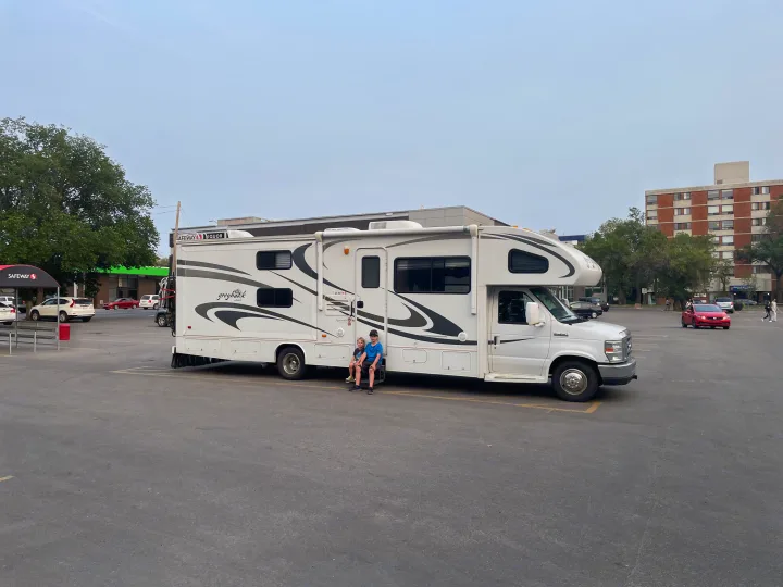 White RV rental in a commercial lot, two boys seated on the steps, with Safeway and other buildings in the background.. Jayco Greyhawk 2010