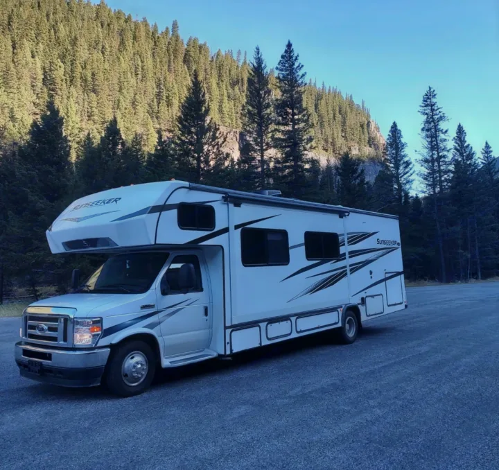 White Sunseeker RV rental parked on a gravel lot, with towering green pine forests and a clear blue sky.. Forest River Sunseeker Classic 2023