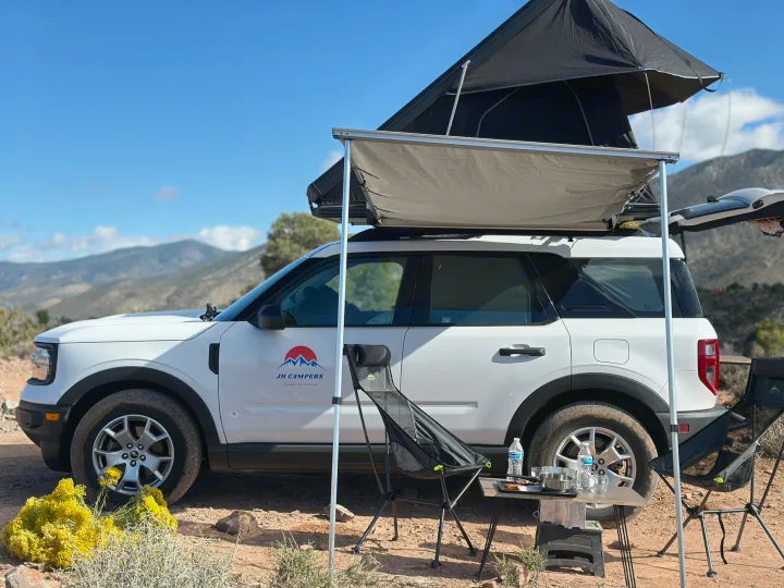 White SUV with rooftop tent & awning for RV rental. Camp setup in scenic mountains, perfect for outdoors camping.. Ford Bronco Sport 2022