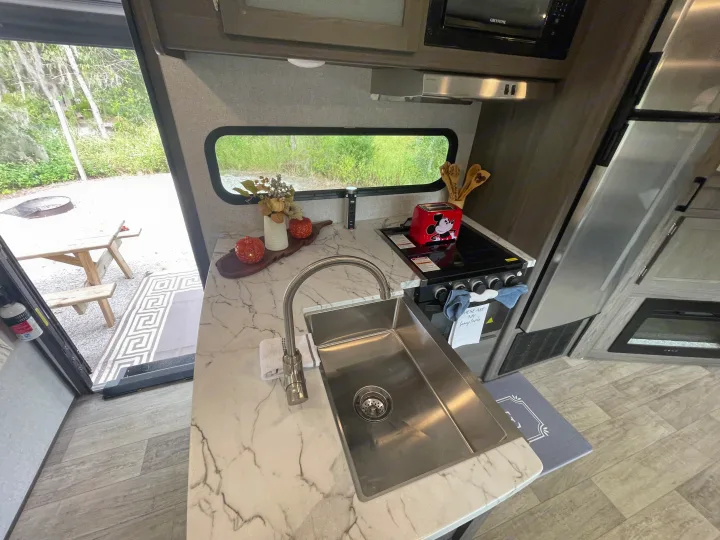 Modern RV rental kitchen with sink, stove, microwave, and festive decor, offering a view of the campsite and picnic area.. Dutchmen Kodiak Ultra Lite 2021