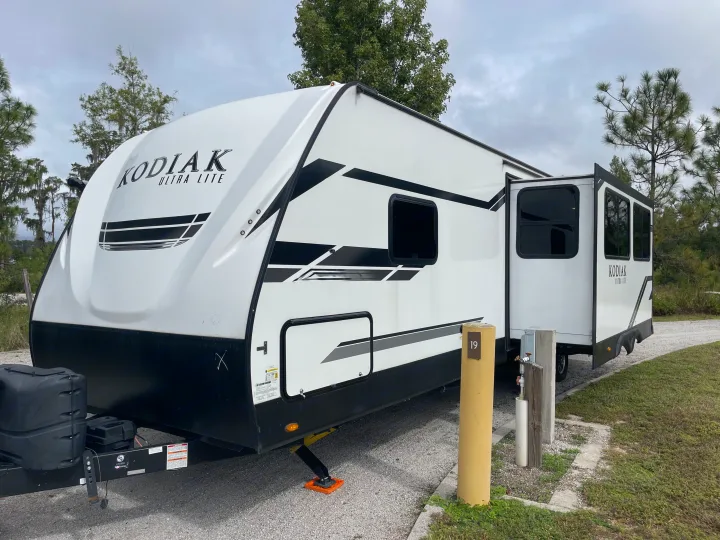 A white KODIAK Ultra Lite travel trailer, a spacious RV rental, parked at an outdoor campsite with slide-out extended. Site 19.. Dutchmen Kodiak Ultra Lite 2021