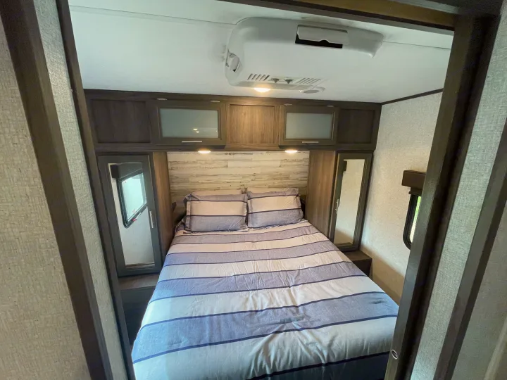 Inside an RV rental: a cozy bedroom with striped bedding, mirrored closets, overhead cabinets, and AC.. Dutchmen Kodiak Ultra Lite 2021