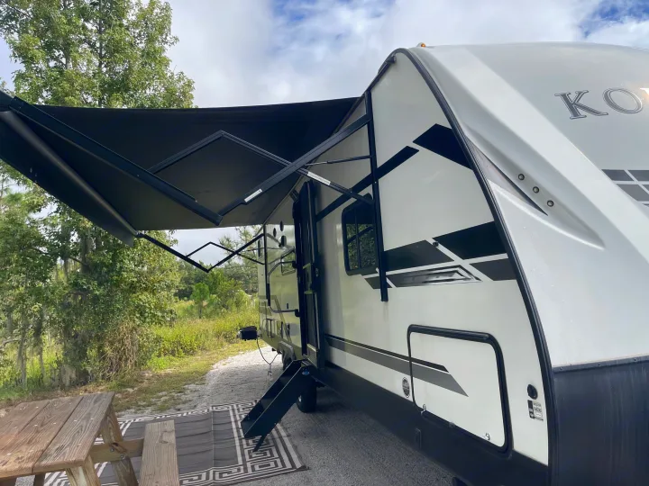 An rv rental with a black awning extended, parked on gravel with a picnic table and rug, nestled in a green campsite.. Dutchmen Kodiak Ultra Lite 2021