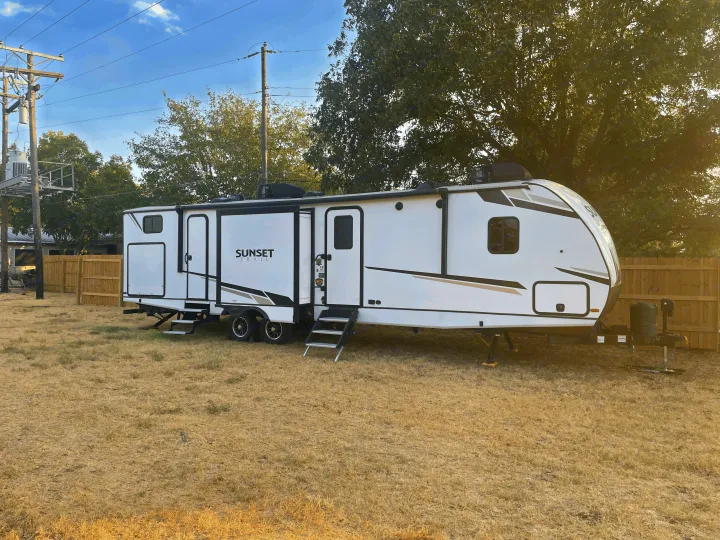 Modern white Sunset Trail travel trailer rv rental parked on a sunny, dry grass lot, with trees and a fence in the background.. Crossroads RV Sunset Trail 2022