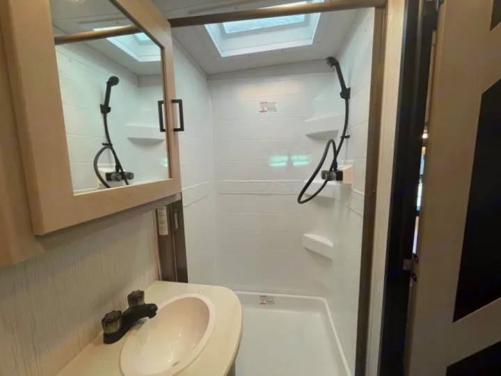 Modern RV rental bathroom: compact sink, mirrored cabinet, and a white shower stall with a handheld shower head and skylight.. Heartland RVs Mallard 2023