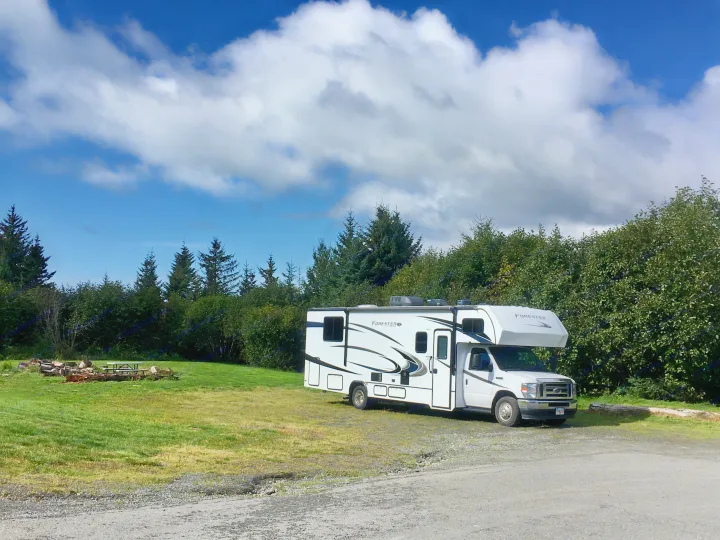 This white Forester RV rental is parked near a green field with a picnic area & tall trees under a cloudy sky.. Forester LE 2022