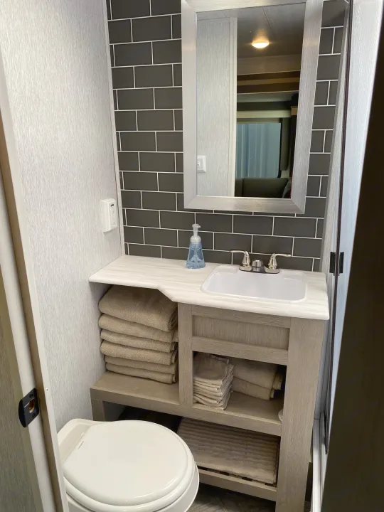 Clean RV rental bathroom featuring a vanity with sink, stacked towels, toilet, and modern dark subway tile backsplash.. Forest River Wildwood 2023