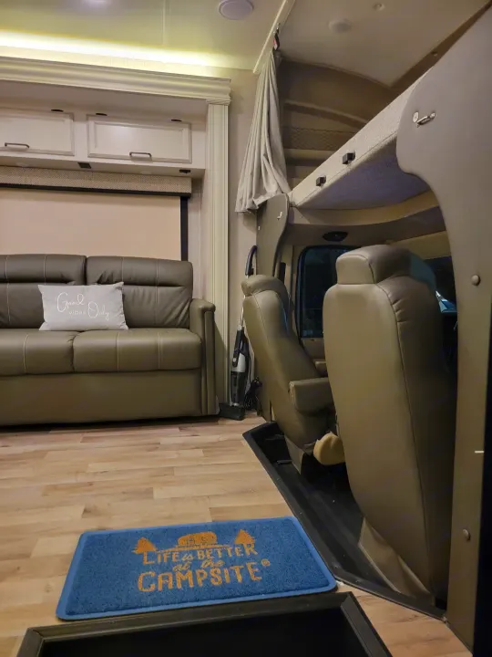 Spacious RV rental interior featuring a cozy sofa, front cab, and overhead bunk, with a welcoming "Campsite" mat.. Jayco Greyhawk 2023