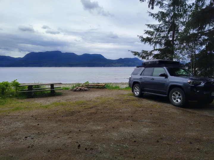 Grey 4Runner with rooftop gear at a scenic lakeside campsite, featuring a picnic table, fire pit, and mountains. RV rental ready!. Toyota 4Runner 2021