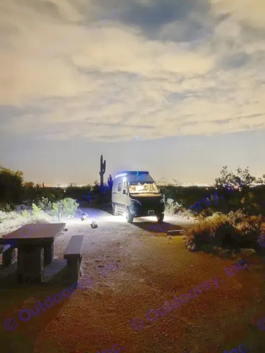 Night desert camping: An RV rental parked under a cloudy sky, with a saguaro and picnic table nearby, city lights in distance.. Winnebago Revel 2024