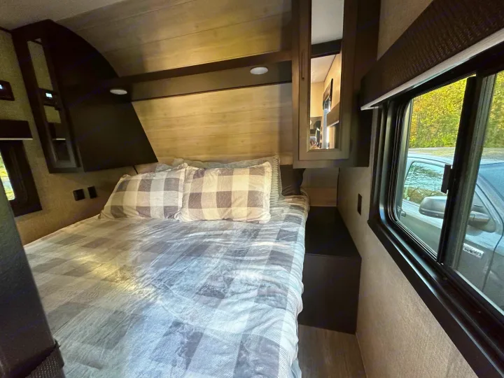 Cozy RV rental bedroom featuring a full bed with plaid bedding, overhead storage, and a window view to nature.. Jayco Jay Flight SLX 2022