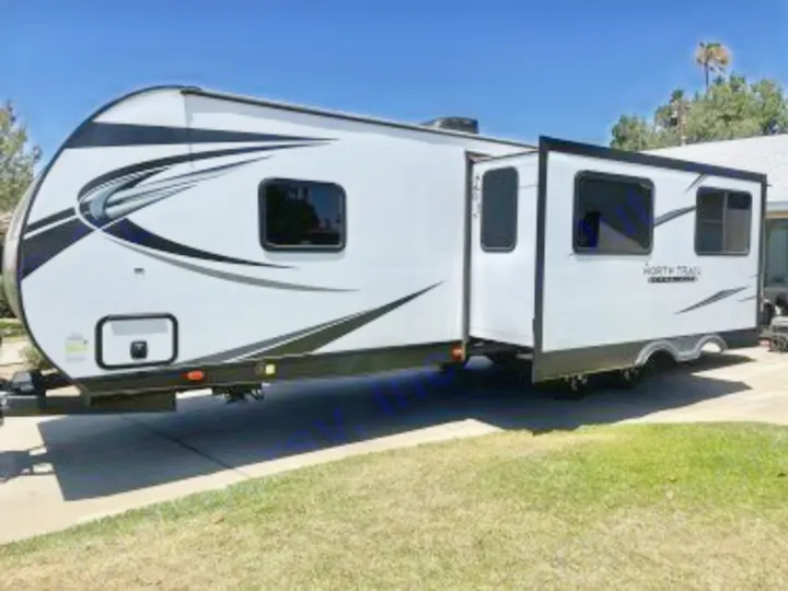 White travel trailer RV with black accents & extended slide-out, parked on a driveway. Great for an RV rental road trip!. Heartland RVs North Trail 2021