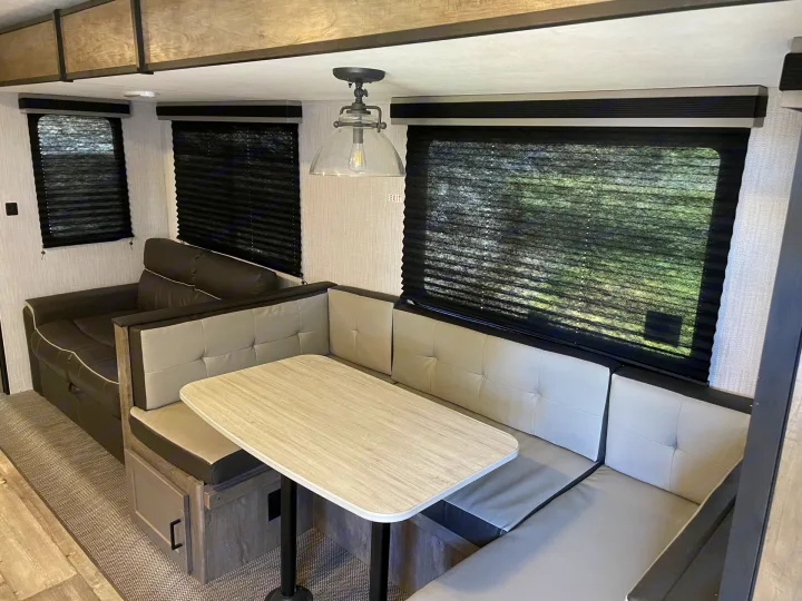 Inside an RV rental: a modern dinette with light gray booth seating and a table, plus a cozy dark sofa and windows.. Heartland RVs North Trail 2021