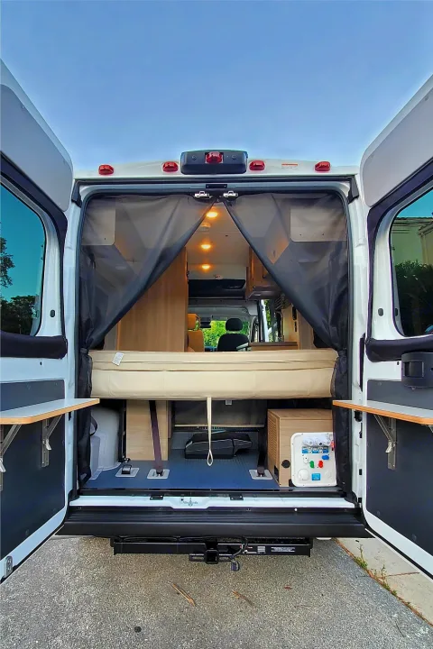 Open RV rental rear view: a cozy bed, accessible storage, and utility panel visible through wide-open doors.. Winnebago Solis 2023
