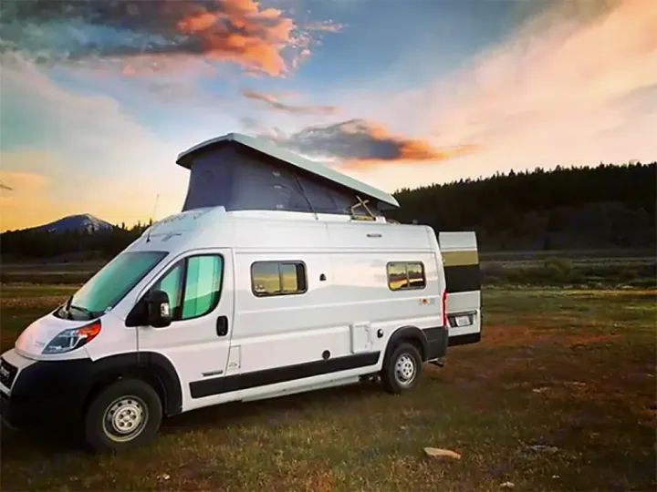 White pop-top RV rental in a scenic field, showcasing mountain views and a vibrant sunset sky. Perfect for camping!. Winnebago Solis 2023