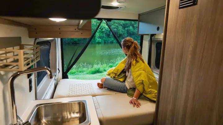 An RV rental interior shows a woman relaxing on a bed, enjoying a beautiful lakeside view through the open rear.. Winnebago Solis 2023