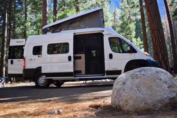 White Solis camper van rv rental with open doors & pop-top in a sunlit forest. Perfect for nature trips.. Winnebago Solis 2023