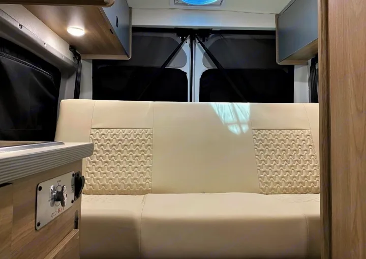 Clean RV rental interior: A cream bench seat with textured details, overhead lights & cabinets, & privacy-covered rear windows.. Winnebago Solis 2023