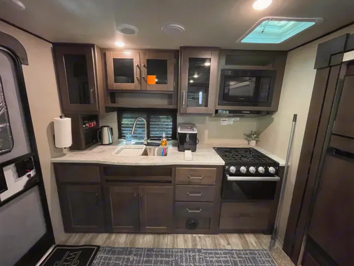 Inside an RV rental: a modern kitchen with dark cabinets, a sink, gas stove, microwave, and ice maker.. Grand Design Transcend Xplor 2022