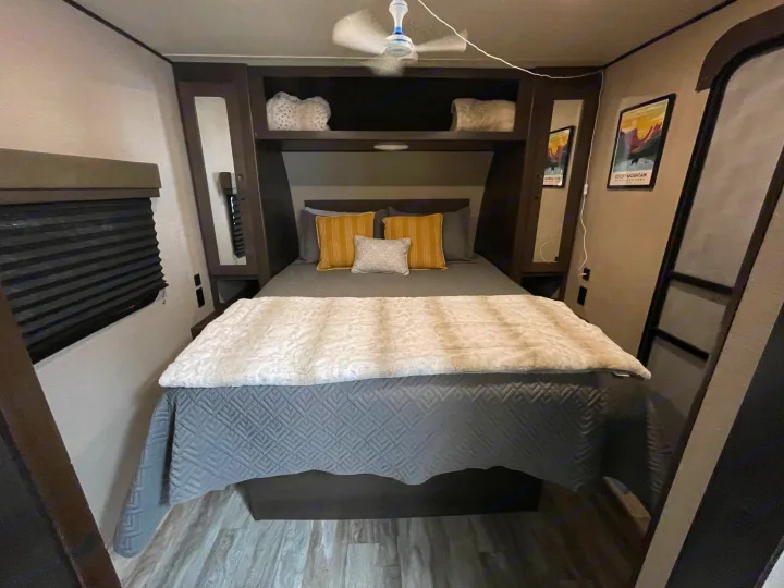 Explore this cozy RV rental bedroom! Featuring a bed with gray bedding, built-in shelves, ceiling fan, and a Rocky Mountain art print.. Grand Design Transcend Xplor 2022