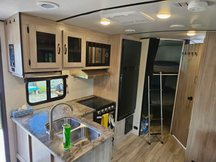 View of an RV rental interior: light wood kitchen with sink, stove, microwave, fridge, and bunk beds with ladder.. Coachmen Northern Spirit 2454BH 2022