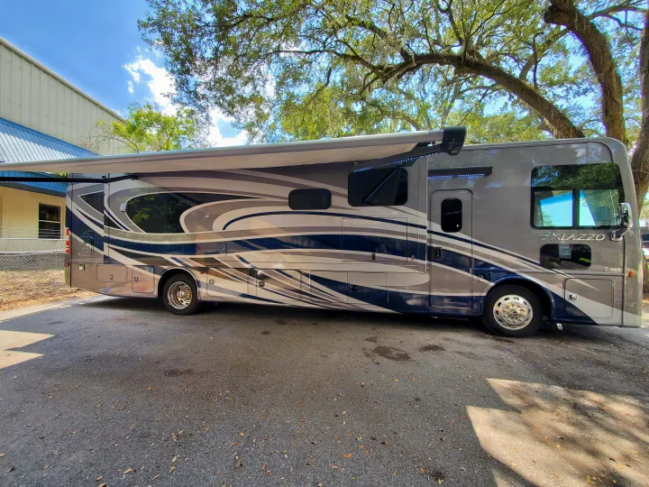 This large Palazzo RV rental, featuring an extended awning with LED lights and a stylish blue/grey design, is parked outdoors.. Thor Palazzo 2022