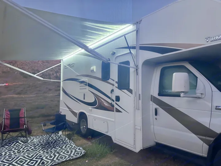 This RV rental offers a perfect evening retreat. A Class C motorhome with a bright LED-lit awning over chairs and a mat.. Thor Freedom Elite 2016
