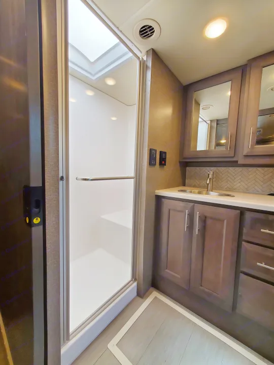 Modern RV rental bathroom: glass shower, skylight, vanity with sink, mirror, dark cabinets, and recessed lighting.. Thor Palazzo 2022