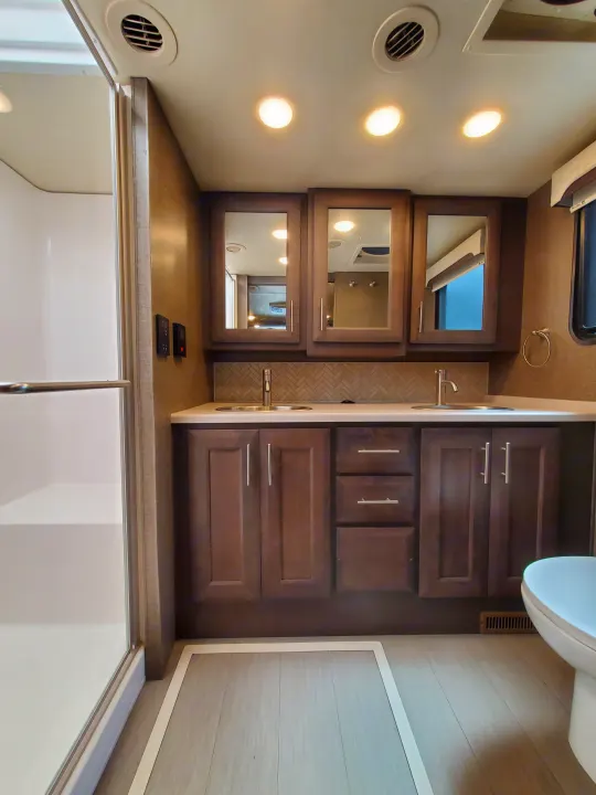 Modern RV rental bathroom: double vanity with two sinks, shower, toilet, and dark wood cabinetry under bright lights.. Thor Palazzo 2022