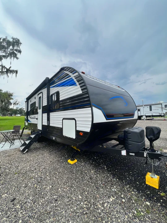 This modern black, white, and blue travel trailer, perfect for an rv rental, is set up at a scenic campsite with a table and chairs.. Keystone Prowler 2021