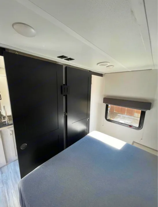 RV rental interior: bedroom with bed, black sliding doors, window, and kitchen area in the background.. Forest River Tracer 2021