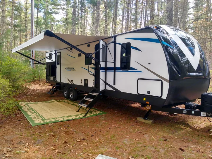 RV rental parked in a sunny, wooded campsite with an extended awning and outdoor mat. Ideal for camping adventures.. Embrace EL310 2021