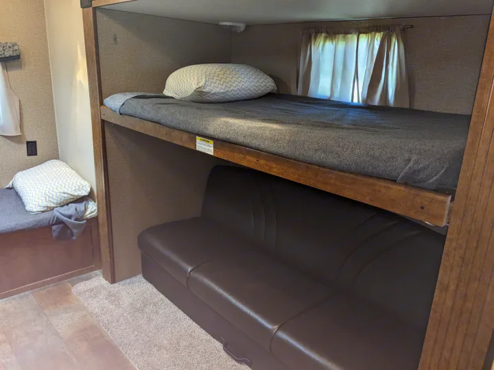 Inside an rv rental, showing bunk beds with gray blankets and a patterned pillow above a brown sofa. Perfect for family travel.. Crossroads RV Z-1 2016