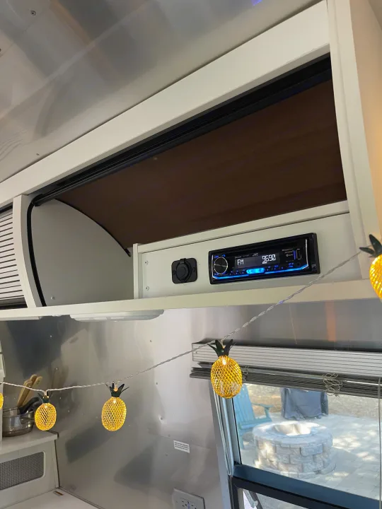 Cozy RV rental interior featuring an entertainment system, power outlet, and overhead storage. Festive pineapple lights adorn the space, with a patio and fire pit visible outside.. Airstream Bambi 2020